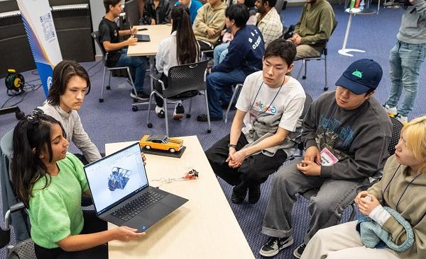 一位身穿问候衬衫、黑发的年轻女子向坐在桌子对面的三名年轻亚洲男子展示了她笔记本电脑上的原型图.  她的男性伴侣坐在她旁边，他面前的桌子上放着一辆橙色的原型车.